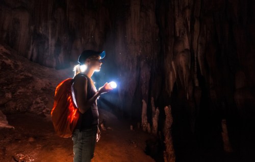 A person with a backpack holds a flashlight, exploring a dark cave with stalactites and a mysterious atmosphere.
