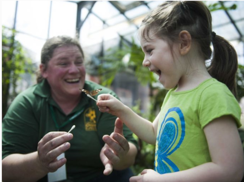 A woman and a girl smile as a butterfly lands on the girl's finger in a greenhouse setting.