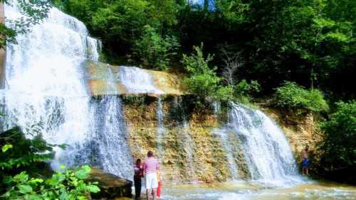 A scenic waterfall surrounded by lush greenery, with two people standing near the water's edge.