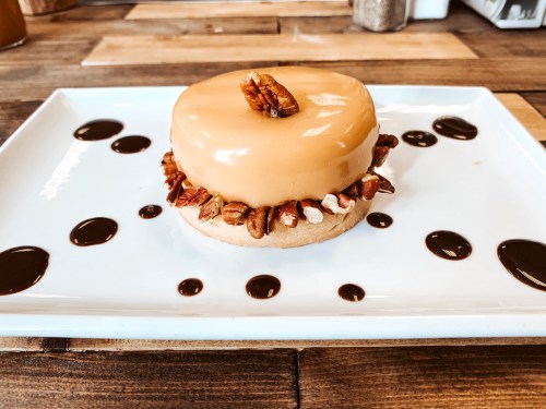 A round dessert topped with caramel glaze and pecans, surrounded by chocolate sauce on a white plate.
