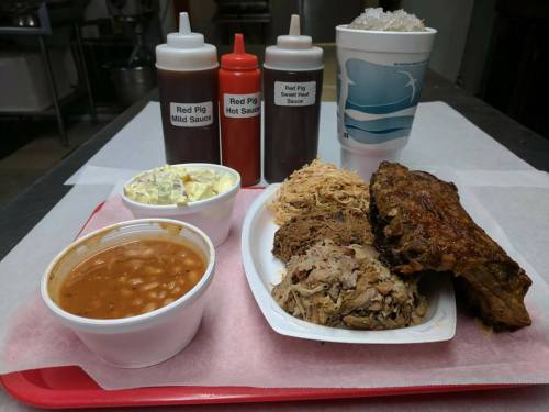 A tray with pulled pork, ribs, coleslaw, baked beans, sauces, and a drink, all served on a red tray.