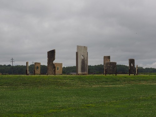 A field with abstract stone structures standing against a cloudy sky, surrounded by green grass.