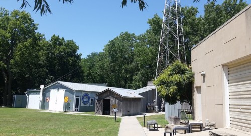 A sunny outdoor scene featuring several buildings and a tall radio tower, surrounded by trees and green grass.