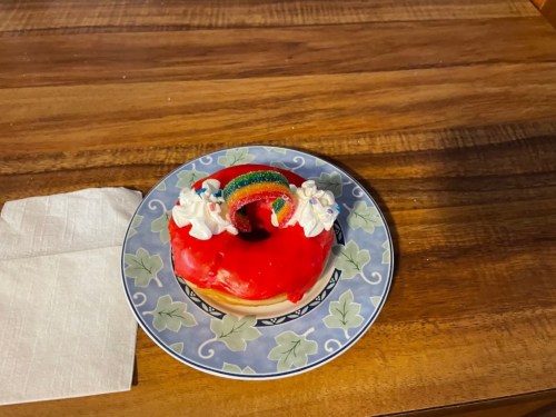 A colorful donut with red icing, topped with whipped cream and a rainbow decoration, on a decorative plate.
