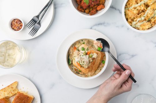 A hand holds a spoon over a bowl of creamy chicken and vegetables, with sides of cornbread and seasoned potatoes.