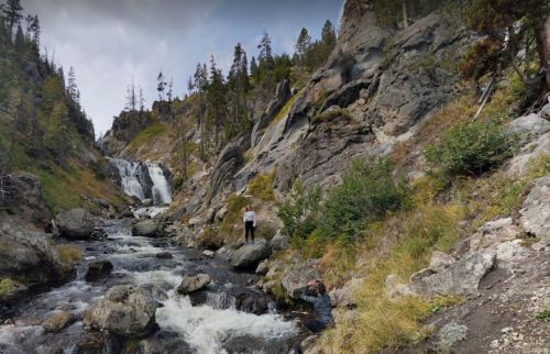 A person stands near a rocky riverbank with a waterfall cascading in the background, surrounded by trees and mountains.