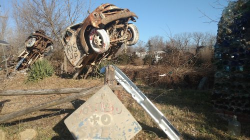 A rusted car is propped up on its rear wheels in a field, with a weathered sign and a bottle wall in the background.