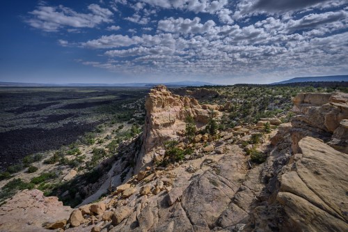 A rocky landscape with layered cliffs, scattered vegetation, and a vast, cloud-filled sky above a dark lava field.