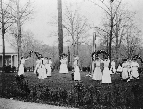 A group of women in white dresses holding floral wreaths, gathered in a park-like setting with trees in the background.