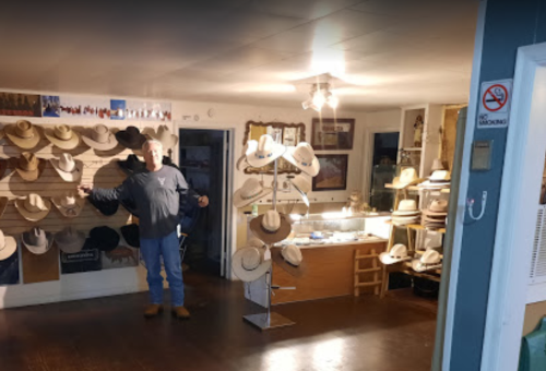 A man stands in a hat shop surrounded by various hats displayed on walls and racks.