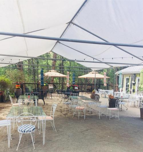 Outdoor seating area with tables and chairs under a large canopy, surrounded by greenery and decorative lights.
