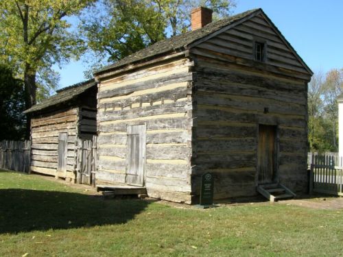 A rustic log cabin with two sections, featuring wooden doors and a grassy area surrounded by trees.