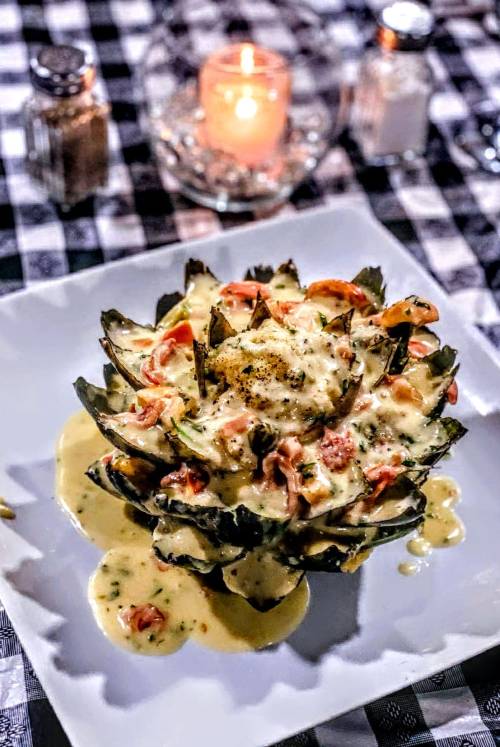 A beautifully plated artichoke topped with creamy sauce, shrimp, and diced tomatoes, set on a checkered tablecloth.
