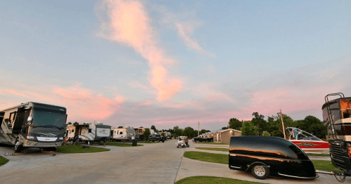 A scenic RV park at sunset, featuring various RVs and a colorful sky with pink and blue hues.