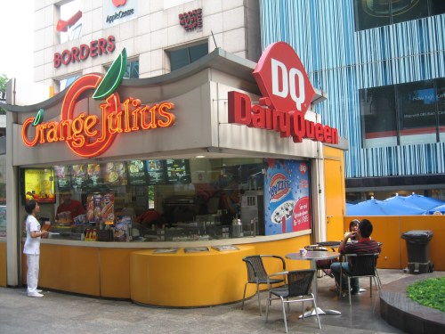 A colorful fast-food stand featuring Orange Julius and Dairy Queen signage, with outdoor seating and customers.
