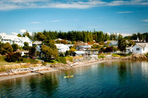 A serene coastal scene with a kayak on calm waters, surrounded by houses and lush greenery under a clear blue sky.