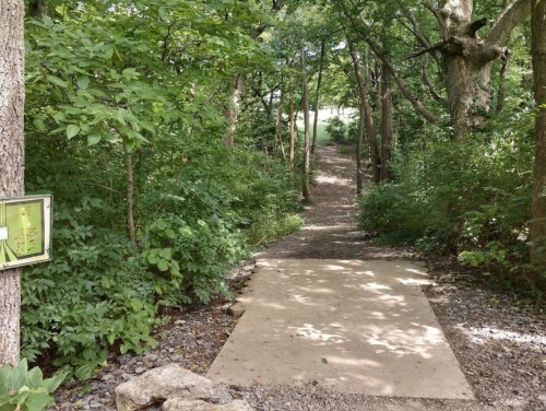 A narrow, tree-lined path leads into a green forest, with a sign on the left and a concrete walkway.