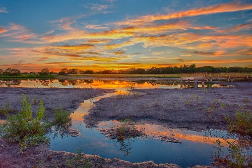 Sunset over a tranquil marsh, with vibrant clouds reflecting in the water and a wooden pier in the background.
