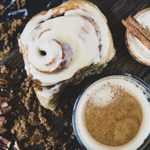 A cinnamon roll with cream frosting beside a glass of coffee, surrounded by brown sugar and cinnamon sticks.