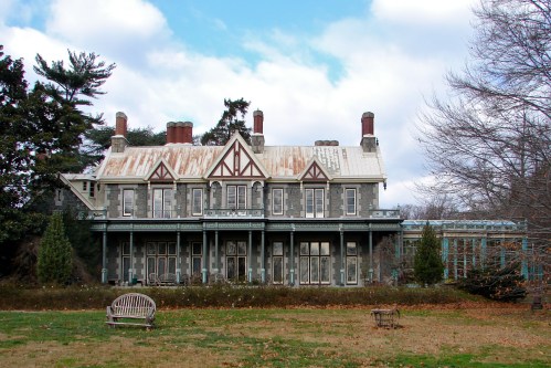 A large, historic mansion with a weathered roof, surrounded by trees and a grassy area, under a cloudy sky.