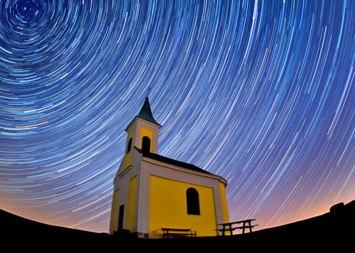 A yellow church under a starry sky, with star trails creating circular patterns around it.