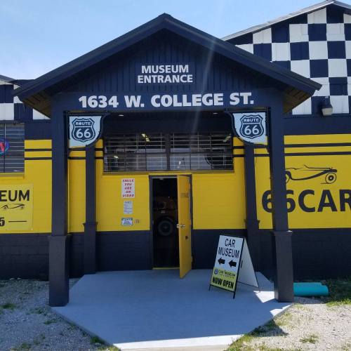 Entrance to a car museum on Route 66, featuring a yellow and black building with a sign indicating it's open.