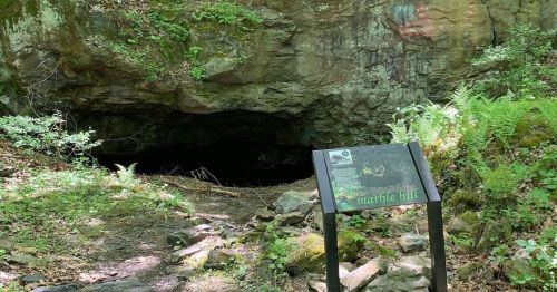 A dark cave entrance surrounded by greenery, with a sign labeled "Marble Hill" in the foreground.