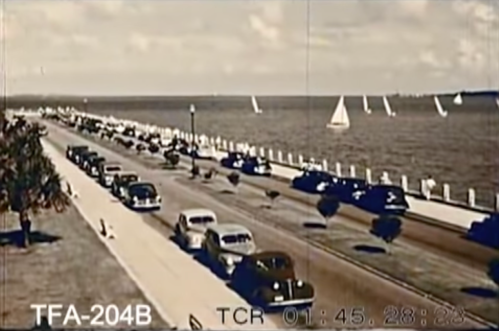 A vintage scene of a waterfront road with cars and sailboats, under a bright sky.