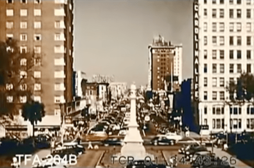 A historic city street scene with cars, buildings, and a central monument, captured in vintage color tones.