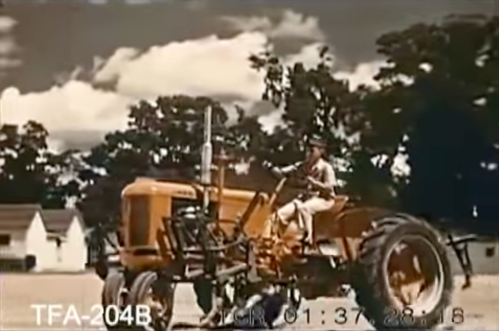 A person operates a yellow tractor in a rural setting with trees and houses in the background under a cloudy sky.