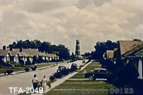 A vintage street scene with houses, a clock tower in the distance, and two people walking along the sidewalk.