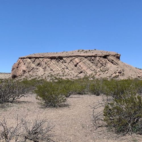A rocky outcrop with layered formations rises above a desert landscape, surrounded by sparse vegetation under a clear blue sky.