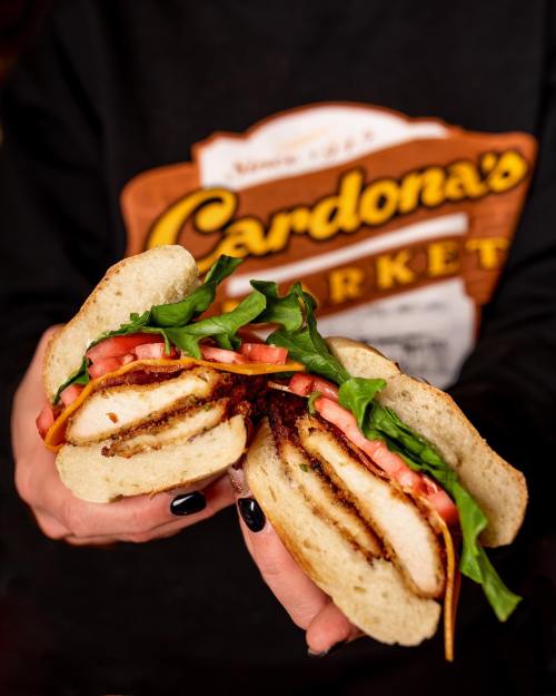 A close-up of a sandwich with crispy chicken, lettuce, and tomato, held by a person against a branded background.