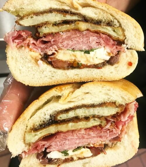 A close-up of a sandwich cut in half, featuring layers of meats, cheese, and fried ingredients between soft bread.