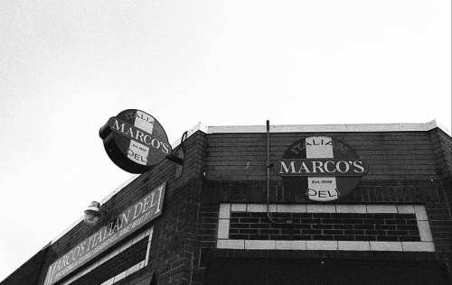 Black and white image of Marco's Italian Deli sign on a brick building against a clear sky.