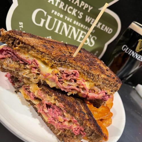 A stacked sandwich with corned beef, cheese, and sauerkraut, served with a side of chips and a Guinness sign in the background.