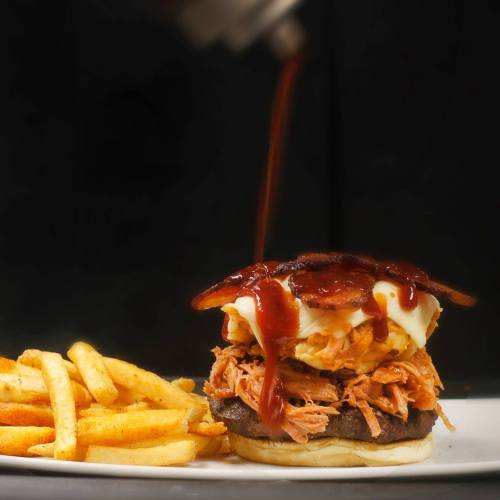 A stacked burger with pulled pork, bacon, and sauce, served with a side of golden fries on a white plate.