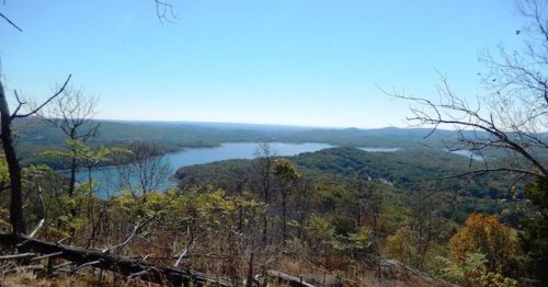A panoramic view of a serene lake surrounded by lush green trees and rolling hills under a clear blue sky.