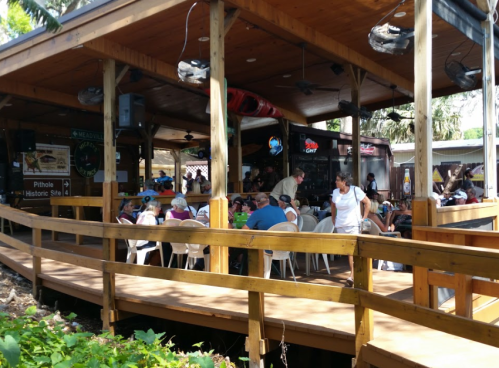 A lively outdoor dining area with people seated at tables under a wooden structure, surrounded by greenery.