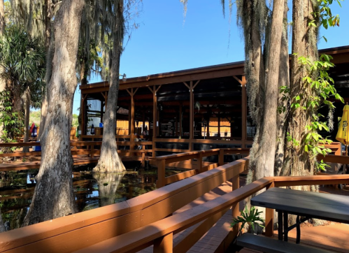 A wooden deck surrounds a restaurant by a calm waterway, with trees draped in Spanish moss under a clear blue sky.