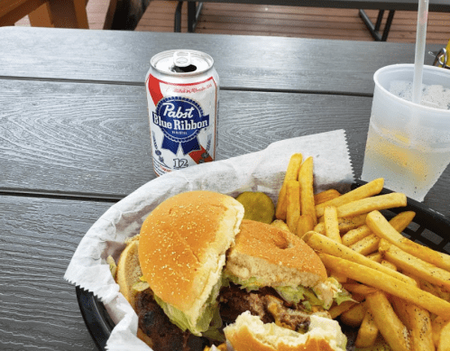 A burger with lettuce and pickles, served with fries and a can of Pabst Blue Ribbon on a wooden table.