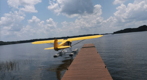 A yellow seaplane is docked at a wooden pier on a calm lake under a partly cloudy sky.
