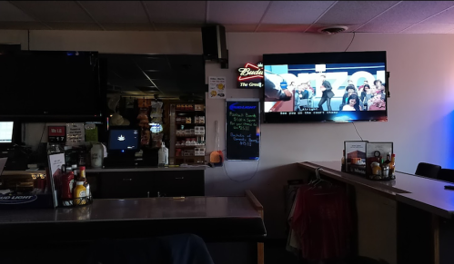 A dimly lit bar with a TV showing a program, shelves of drinks, and a counter with condiments and menus.