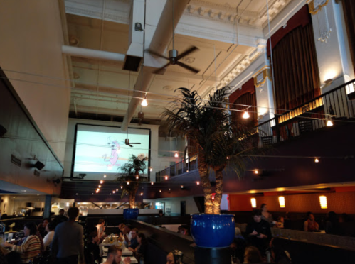 Interior of a restaurant with a large screen showing a cartoon, palm trees, and a lively atmosphere.