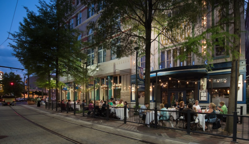 Outdoor dining scene at night, with people seated at tables under trees and string lights along a city street.