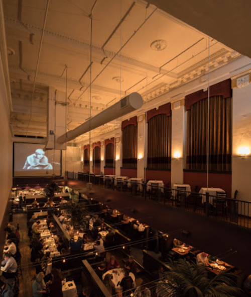 A spacious restaurant with elegant decor, featuring diners at tables and a large screen displaying a film.