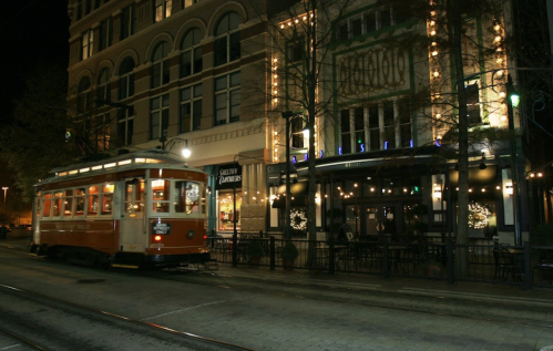 A vintage streetcar passes by a lit-up restaurant on a city street at night.