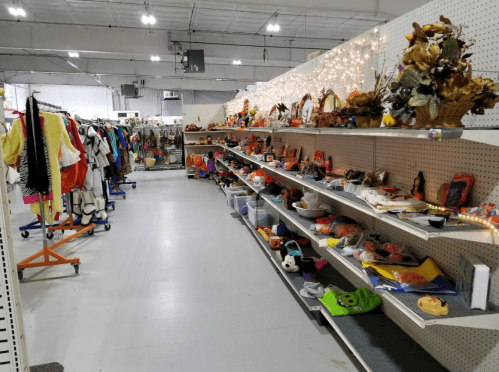 A spacious thrift store aisle with shelves of toys, decorations, and clothing racks on the left side.