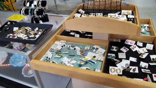 Display of assorted jewelry, including earrings and pins, in wooden trays at a market stall.