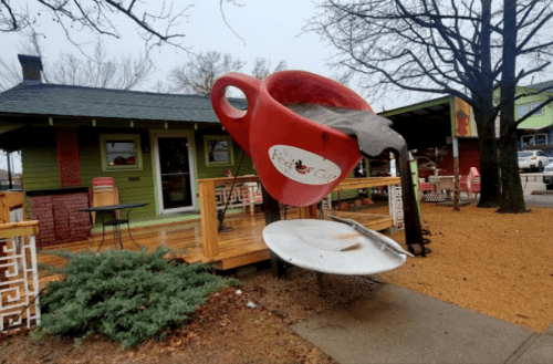 A large red coffee cup sculpture spills into a white saucer outside a colorful café on a rainy day.